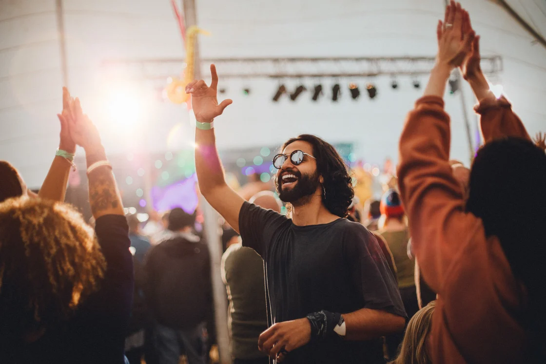 People are dancing in a performance tent at a music festival. The main focus is on a man who is wearing sunglasses and is dancing with a hand in the air.
